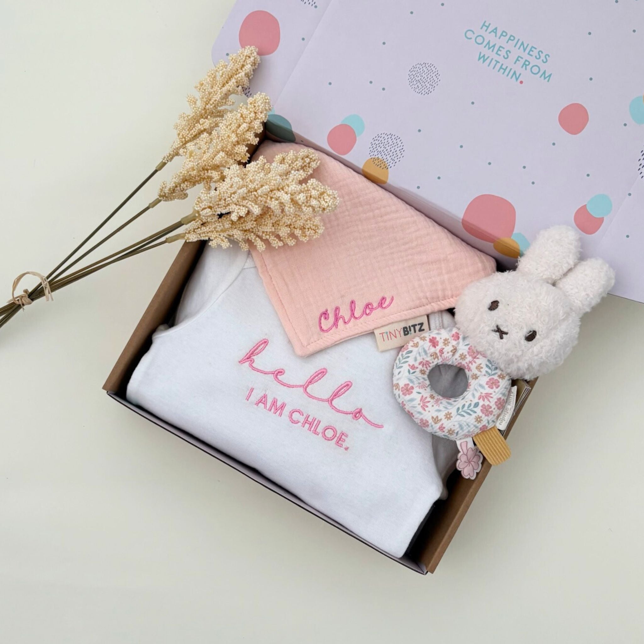 Personalised baby gift set with embroidered onesie, muslin bib, and floral Miffy rattle toy, all neatly packed in a pastel gift box for newborn gifting.